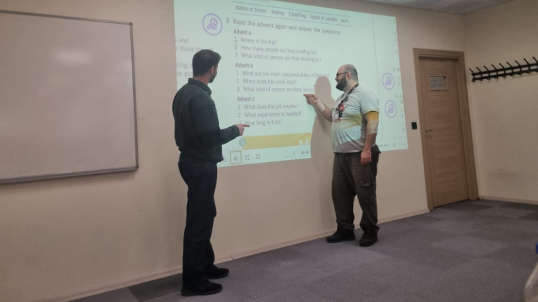 İki kişi sınıfta projeksiyon ekranında iş ilanı sorularını inceliyor, eğitim ortamı.